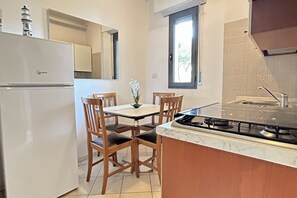 A compact kitchen with a refrigerator, a dining table with chairs, a gas stove, and a window with a view. - San Martino Flexrent - Abissinia Real Estate (Riccione)