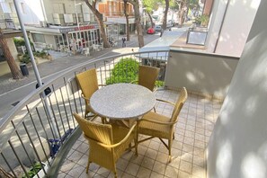 A balcony with a round table and wicker chairs, overlooking a street with shops and pedestrians. - San Martino Flexrent - Abissinia Real Estate (Riccione)