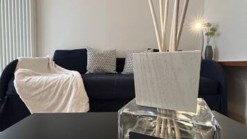 A living room with a dark sofa, patterned cushions, a white throw blanket, and a vase with tall, thin stems.