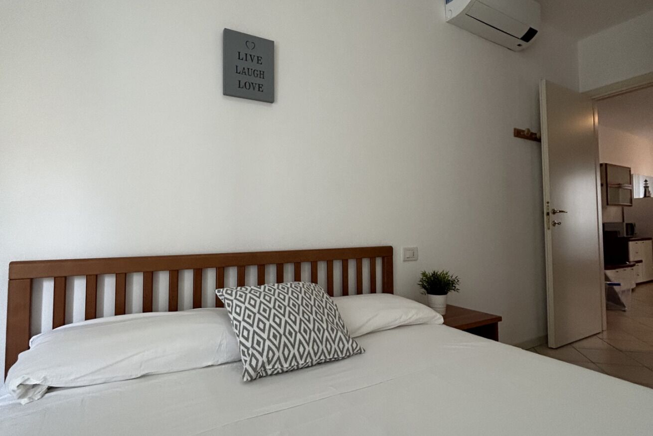 Ein Schlafzimmer mit einem Holzkopfende, einem Bett mit weißer Bettwäsche, einem gemusterten Kissen, einem kleinen Nachttisch mit einer Pflanze und einem an der Wand befestigten Schild.