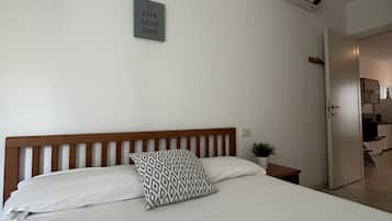 A bedroom with a wooden headboard, a bed with white bedding, a patterned pillow, a small bedside table with a plant, and a wall-mounted sign.