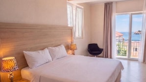 Hotellihuone, jossa on suuri sänky, yöpöytä lampun kanssa, tuoli ja parveke, jolta on näkymä merelle.