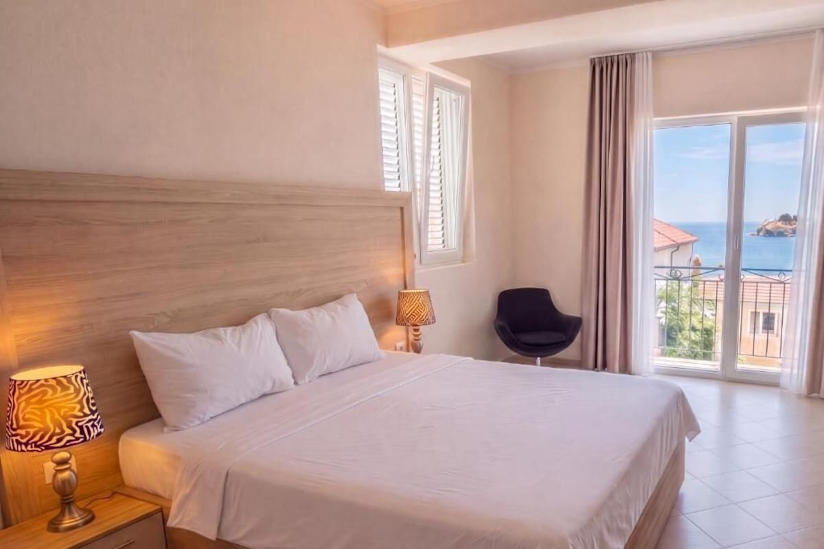 A hotel room with a large bed, a nightstand with a lamp, a chair, and a balcony with a view of the sea.