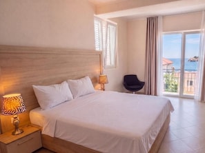 A hotel room with a large bed, a nightstand with a lamp, a chair, and a balcony with a view of the sea. - Casa Viktoria (Sveti Stefan)