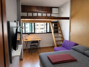 A compact room with a bunk bed, a desk, a chair, a TV, and a window with a view of buildings. - Vrbo Property (Rio de Janeiro)