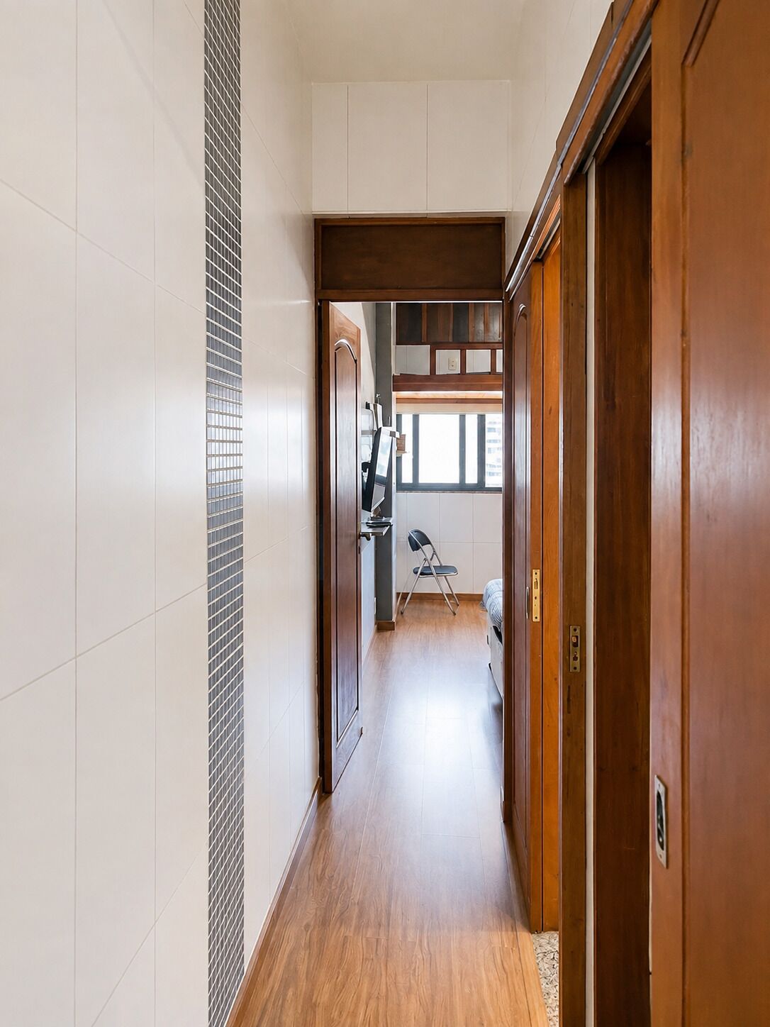 Een smalle gang met houten vloeren, een witte betegelde muur en een houten deur aan de rechterkant.