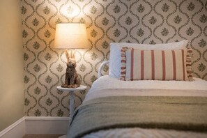 Ein Schlafzimmer mit raumhoher Tapete mit Muster, einem Nachttisch mit Lampe und einem Bett mit gestreiftem Kopfkissen.