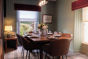 A dining area with a wooden table set for four, surrounded by chairs, with a window view and a lamp on a side table. - Fairytale Scots Baronial Coach House in Ballater, near Balmoral Castle (Ballater)