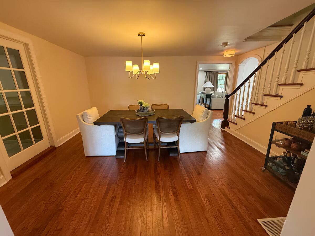 A dining area with a wooden floor, a chandelier, and a staircase in the background.