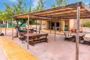 Un patio al aire libre con bancos y mesas de madera, una zona de estar cubierta y un muro de piedra al fondo.