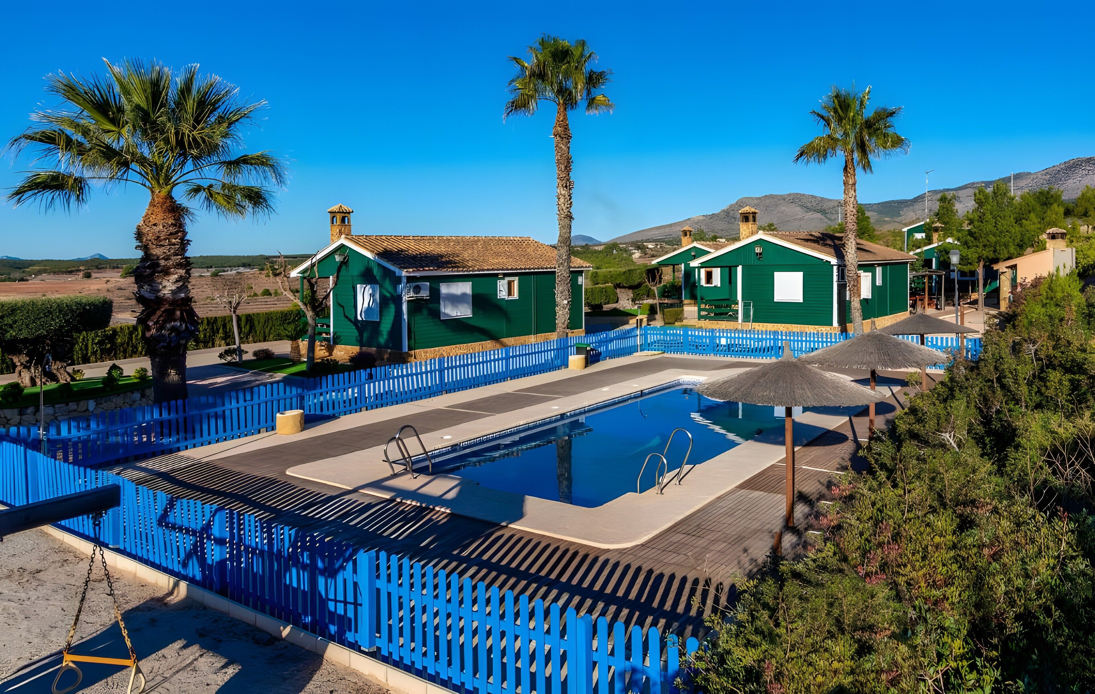 A pool area with a blue fence, green buildings, and palm trees.