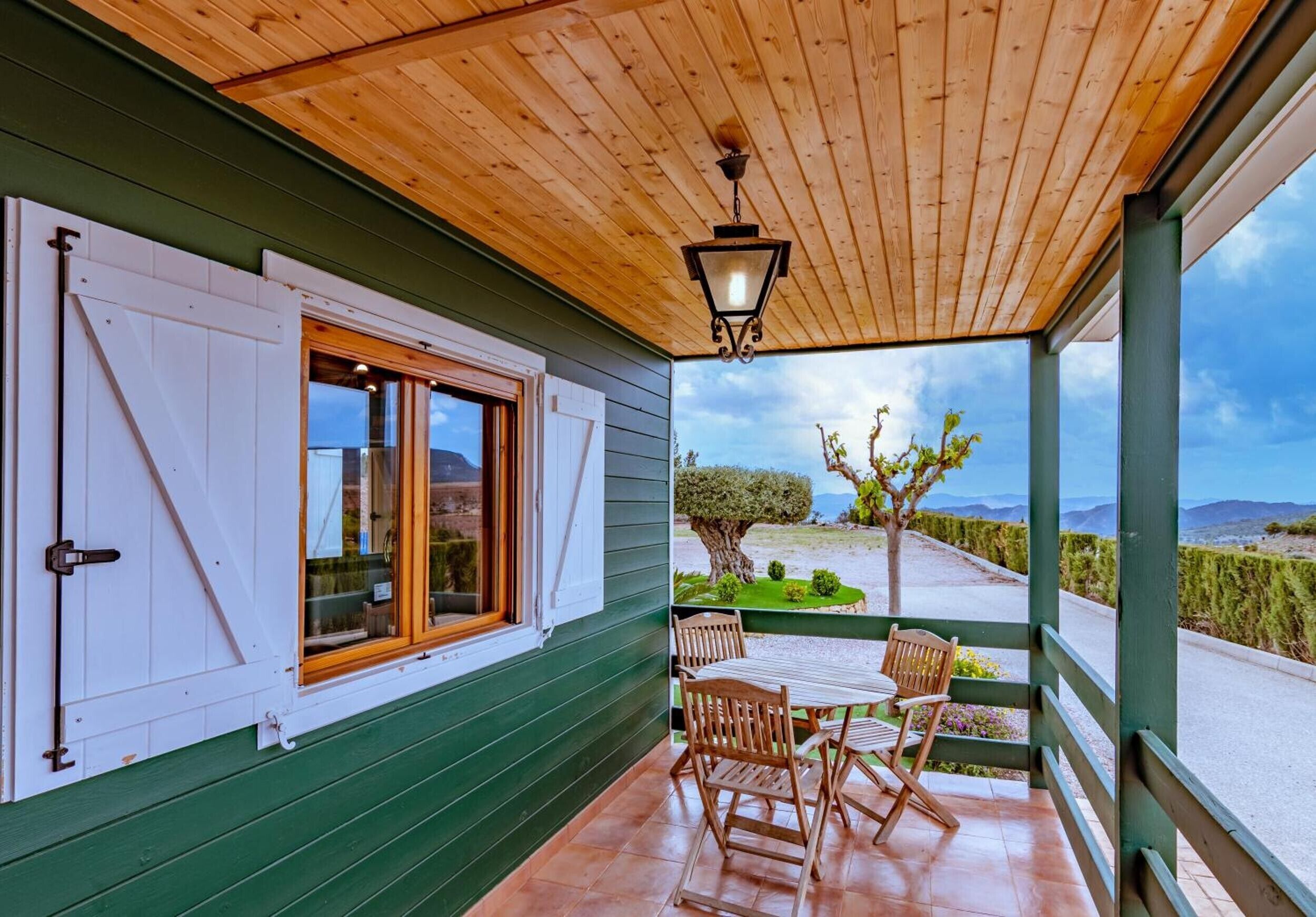 Eine überdachte Veranda mit Holzboden, einem Tisch mit Stühlen und Blick auf eine Landschaft mit Bäumen und Bergen.