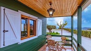 Een overdekt terras met houten plafond, een tafel met stoelen en uitzicht op een landschap met bomen en bergen.