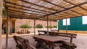 Un patio al aire libre con mesas y bancos de picnic de madera, un área de estar cubierta con techo de bambú y un muro de piedra al fondo.