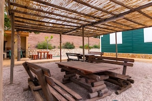 An outdoor patio area with wooden picnic tables and benches, a covered seating area with a bamboo roof, and a stone wall in the background.