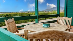 Een houten tafel met een groene blik erop, omringd door houten stoelen, op een balkon met uitzicht op een groen veld en een berg in de verte.