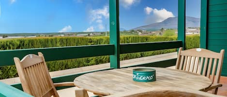 Una mesa de madera con una lata verde, rodeada de sillas de madera, en un balcón con vistas a un campo verde y una montaña a lo lejos.