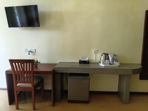 A compact room with a flat-screen TV mounted on the wall, a small table with a chair, a compact refrigerator, and a coffee maker on the table.