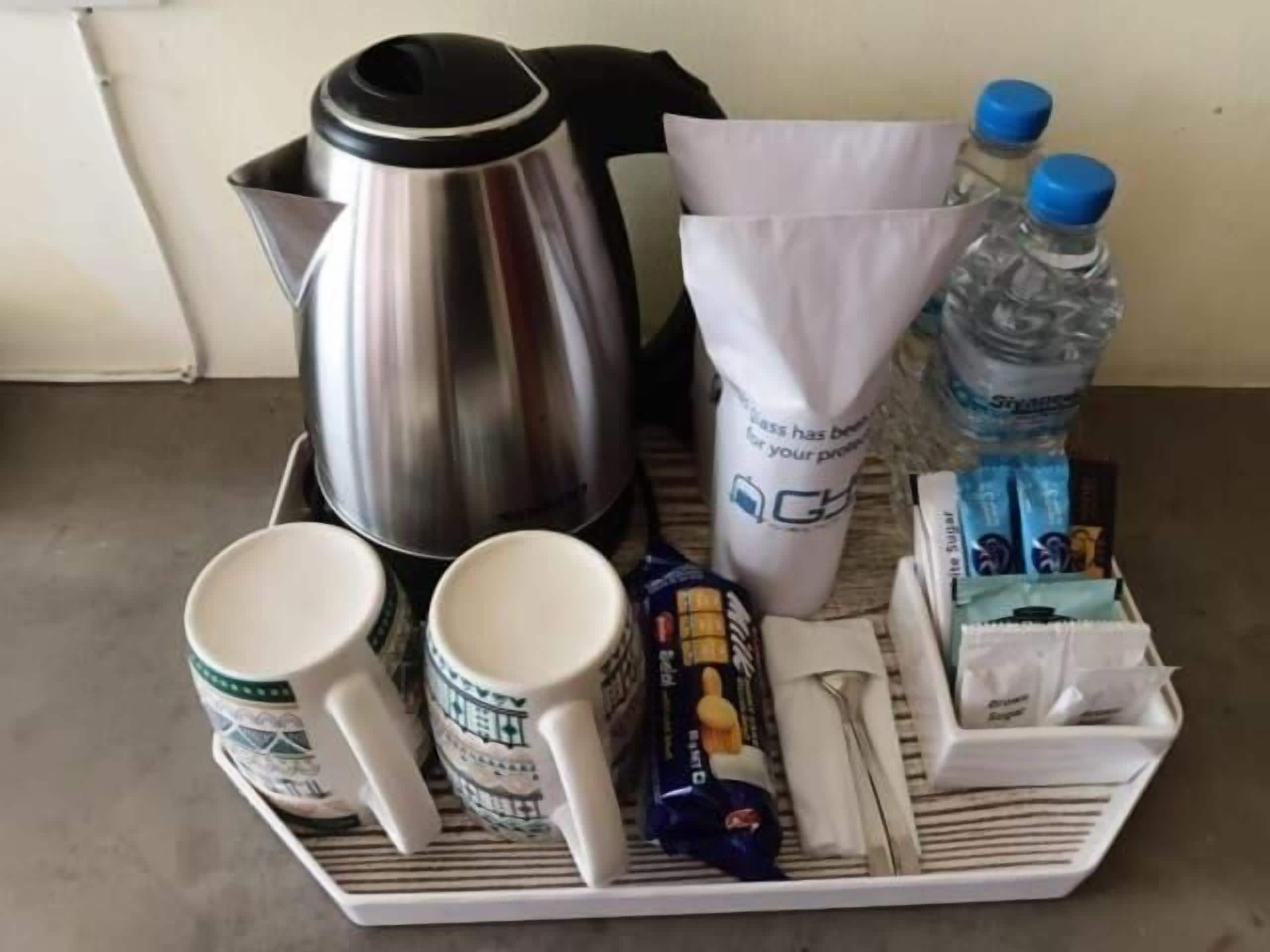 Un vassoio con una caffettiera in acciaio inox, due tazze bianche, una bottiglia d'acqua, un pacchetto di fazzoletti e un piccolo contenitore di snack.