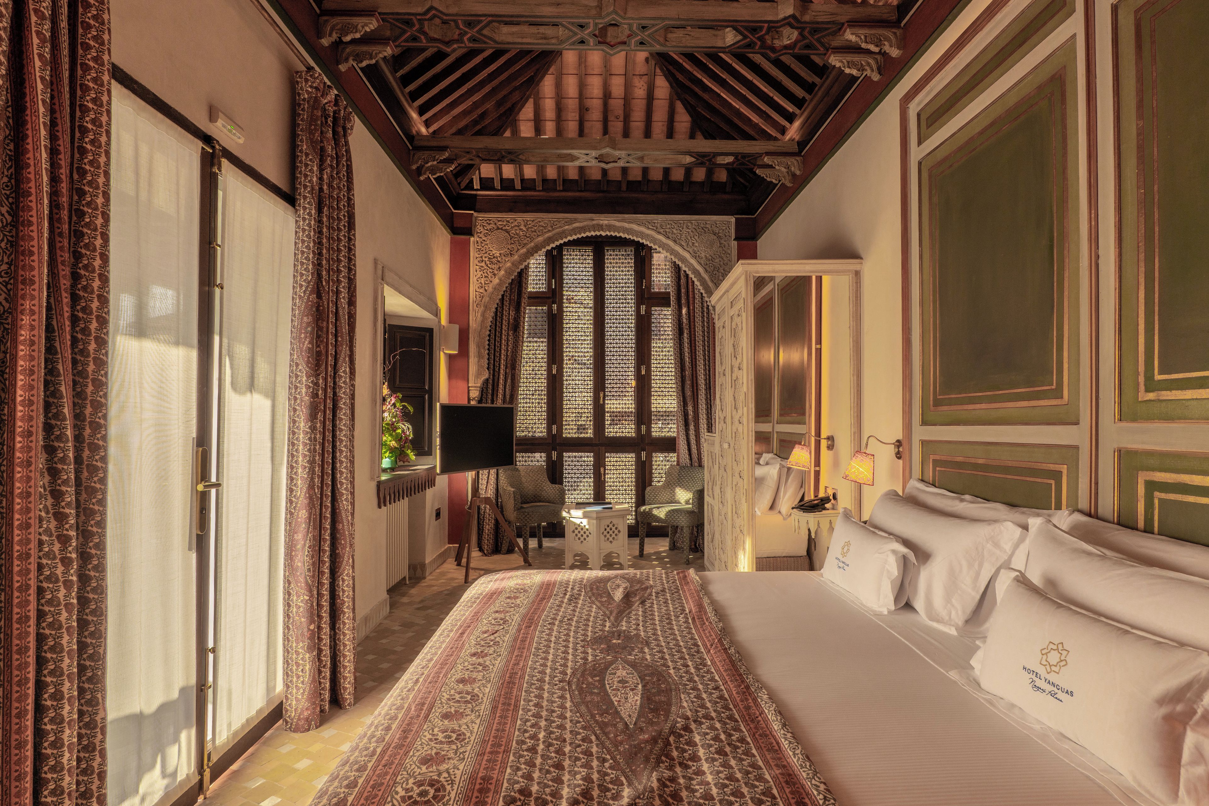 Habitació d'hotel amb un llit gran, zona de seients amb una cadira, escriptori i finestra amb un disseny de celosies detallat.