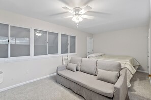 A bedroom with a bed, a sofa, a ceiling fan, and a window with blinds. - Folly Fun House – Just Steps to the Beach (Folly Beach)
