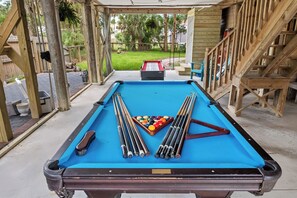 A pool table with balls and cues, surrounded by wooden structures and a grassy yard. - Folly Fun House – Just Steps to the Beach (Folly Beach)