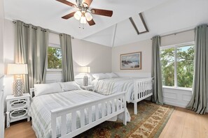 A bedroom with two beds, a ceiling fan, a painting, and a view of trees through the windows. - Folly Fun House – Just Steps to the Beach (Folly Beach)