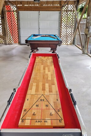 A pool table with a wooden playing surface and a red felt bottom, set in a room with a concrete floor and wooden walls. - Folly Fun House – Just Steps to the Beach (Folly Beach)