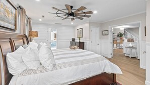 A bedroom with a wooden headboard, a large bed with white bedding, a ceiling fan, and a view into the adjacent dining area. - Folly Fun House – Just Steps to the Beach (Folly Beach)