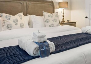 A neatly made bed with a towel and a bottle labeled 'Makeup' on top, a patterned pillow, a lamp on a bedside table, and a dark blue textured blanket.