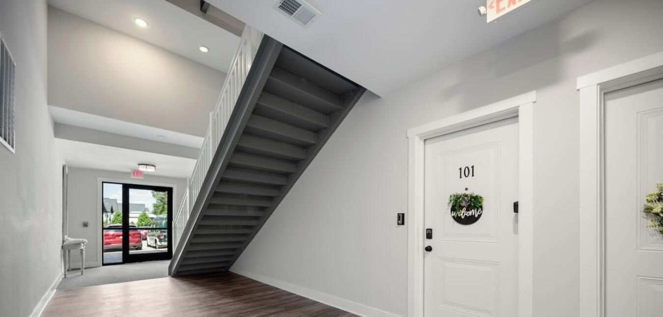 A modern interior with a staircase, a white door labeled '101,' and a view of the outside through glass doors.