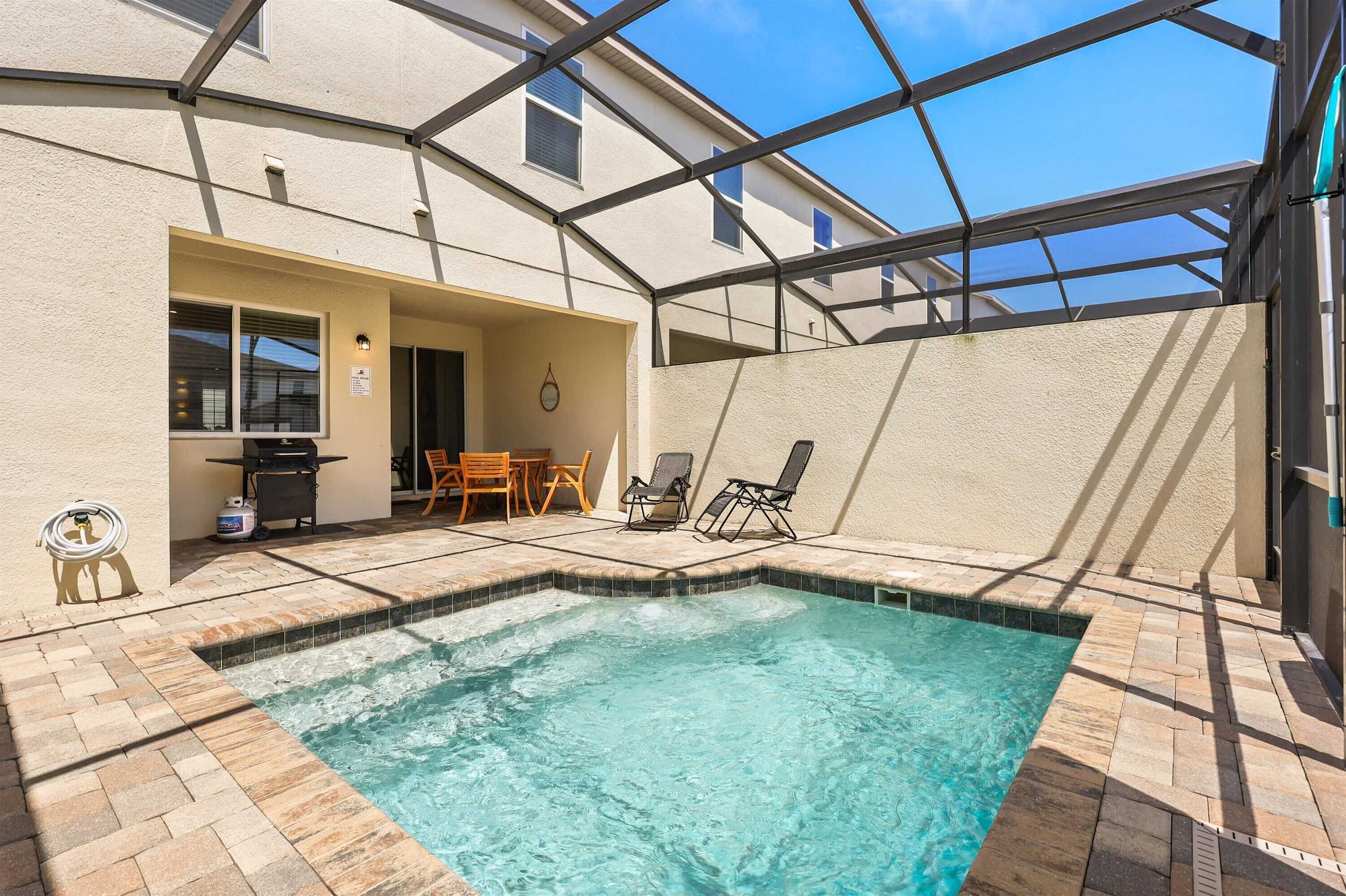 Piscina com água azul cristalina, cercada por um deck revestido e uma área de varanda coberta com cadeiras e mesa.