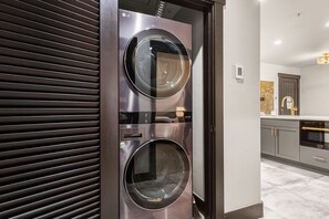 Un moderno lavadero con dos lavadoras y secadoras de acero inoxidable de carga frontal, una puerta corredera, y una zona de cocina con un lavamanos y armarios.