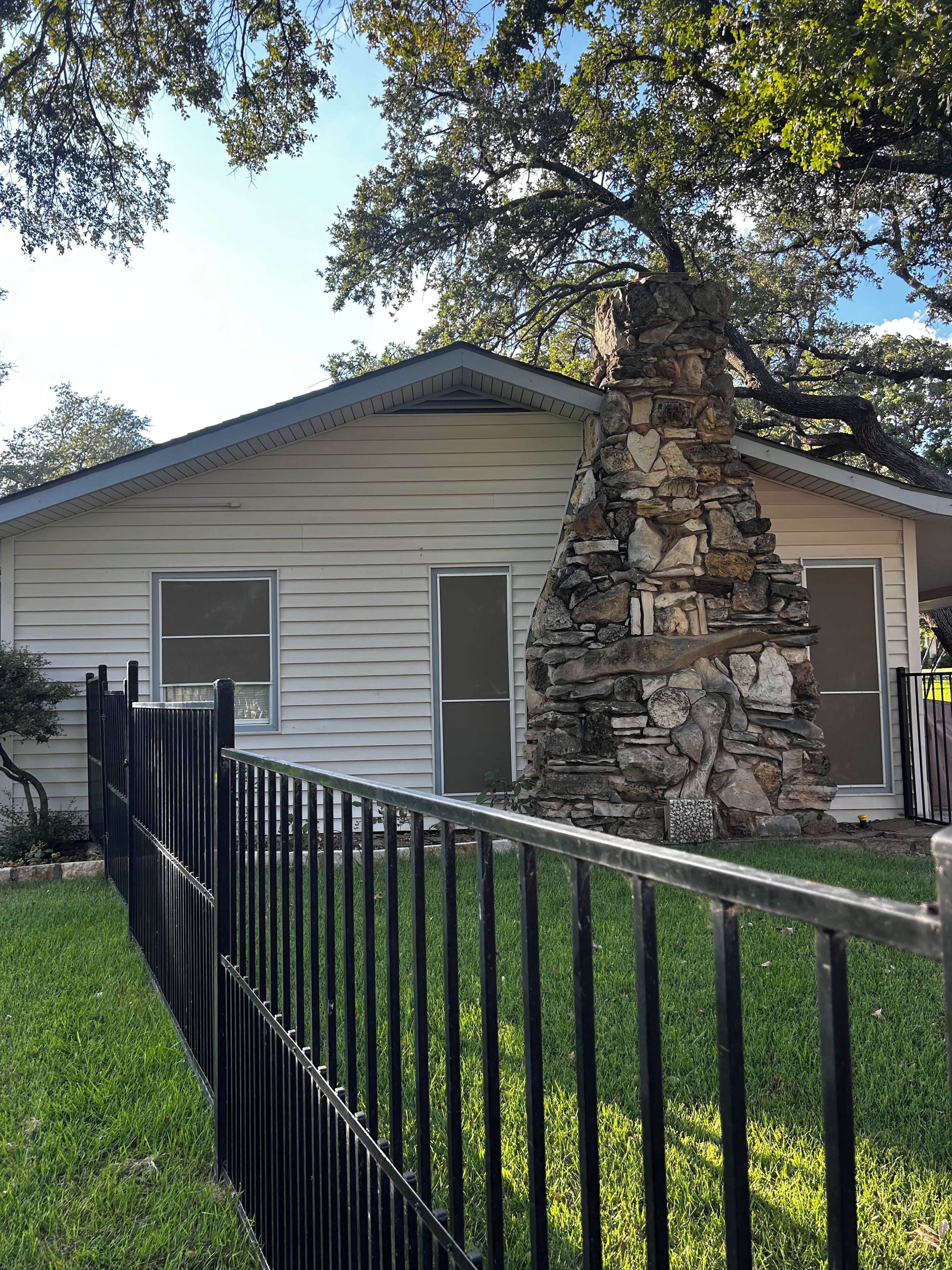 A single-story house with a stone pillar, a black metal fence, and a grassy yard.