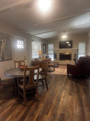 A living room with a fireplace, a dining area with wooden furniture, and a television mounted on the wall. -  Lake Granbury Waterfront Cottage! 2 miles from Downtown with private boat dock! (Granbury)