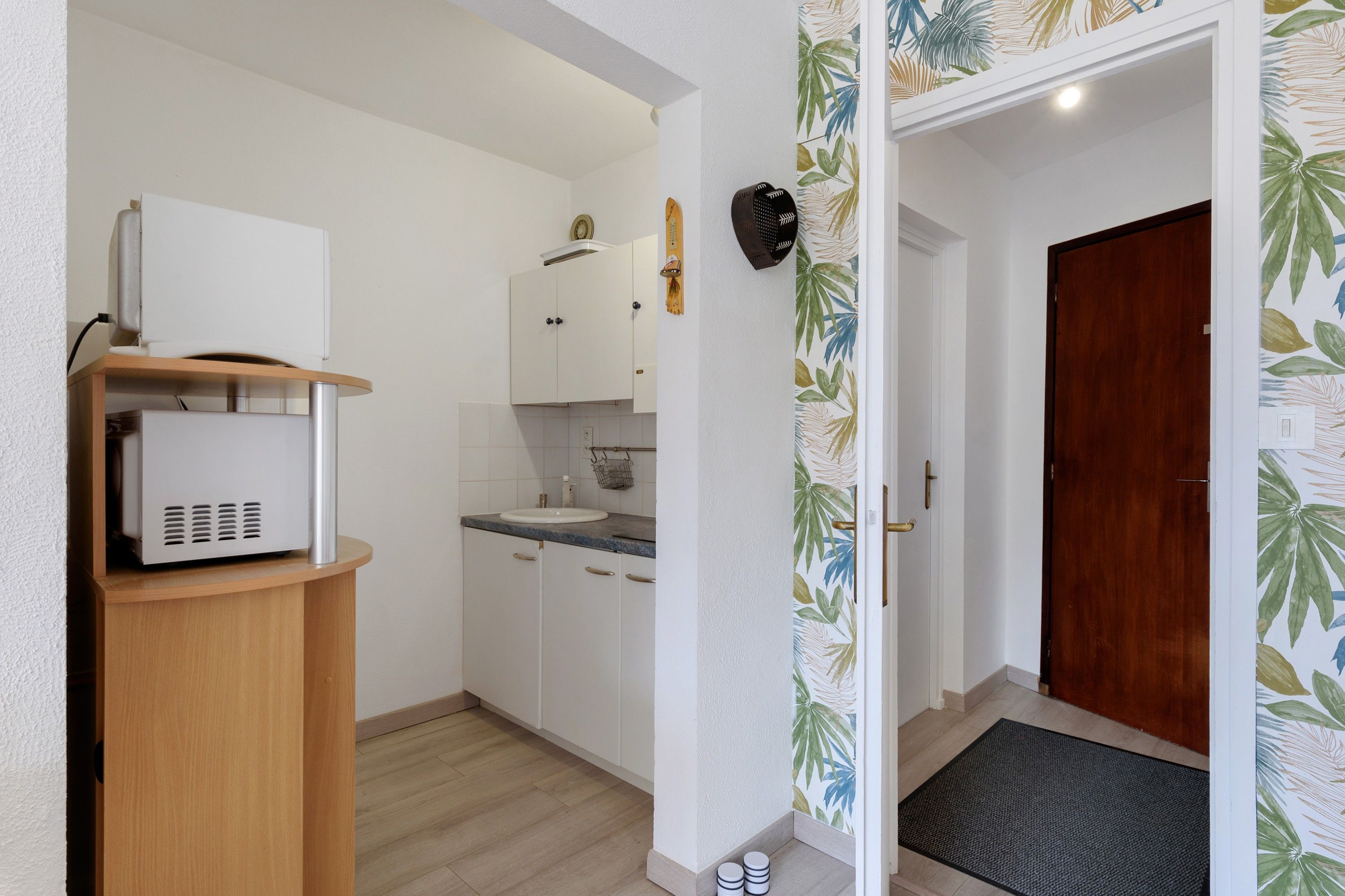 Une cuisine compacte avec des meubles blancs, un four à micro-ondes posé sur un meuble en bois, un évier et une porte ornée d’un motif de feuilles tropicales.