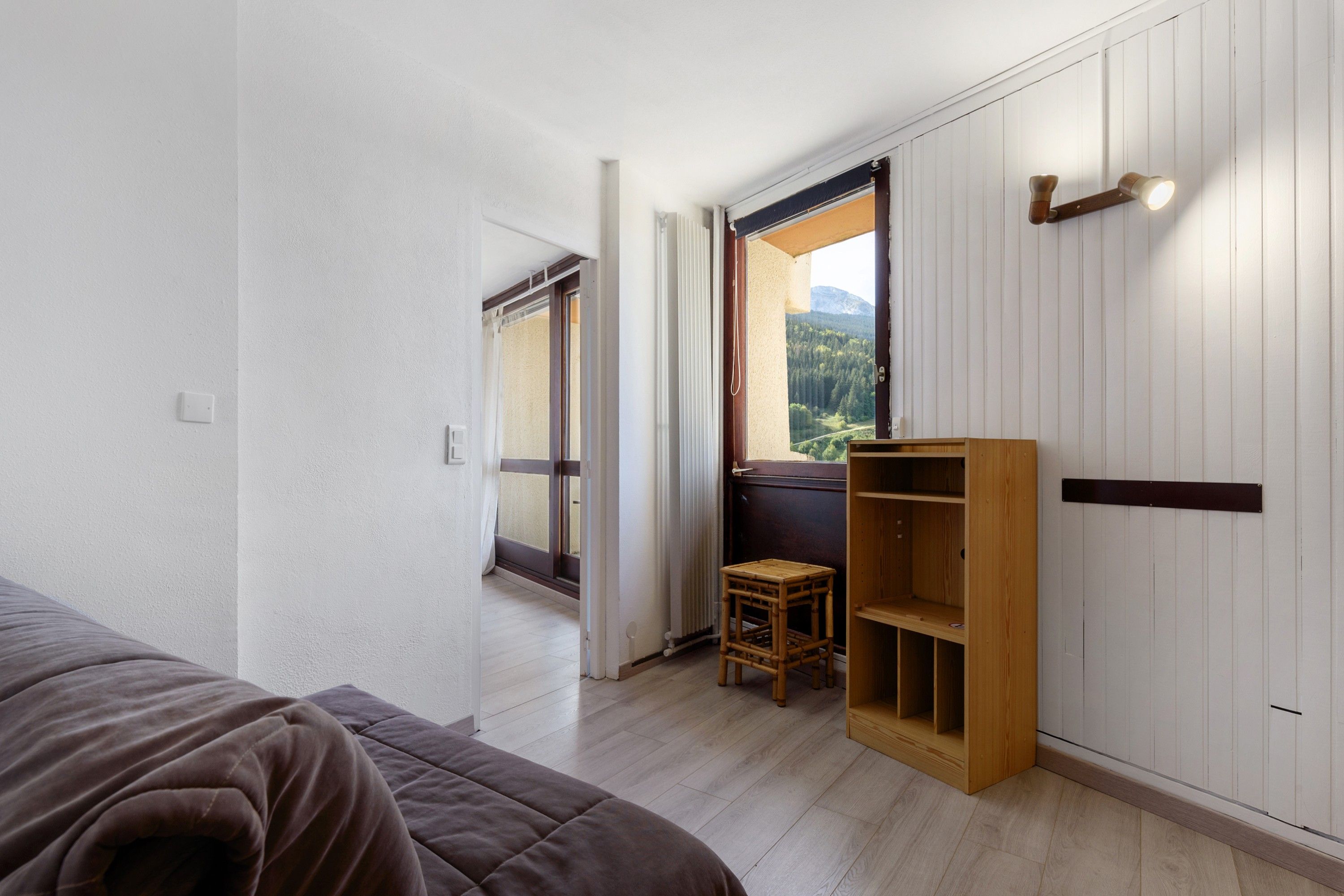 Een slaapkamer met een bed, een houten plank, een klein krukje en een raam met uitzicht op een berg.