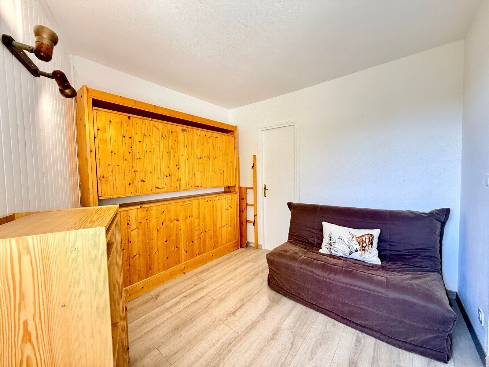 Habitación compacta con un armario de madera, un sofá marrón con un cojín decorativo y una escalera de madera apoyada en la pared.