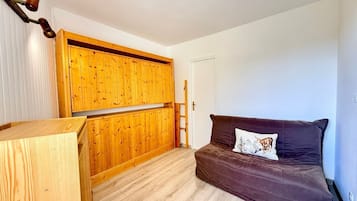 Habitación compacta con un armario de madera, un sofá marrón con un cojín decorativo y una escalera de madera apoyada en la pared.