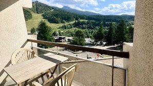Un balcon avec une table et des chaises, donnant sur un paysage verdoyant, avec des arbres et une route.