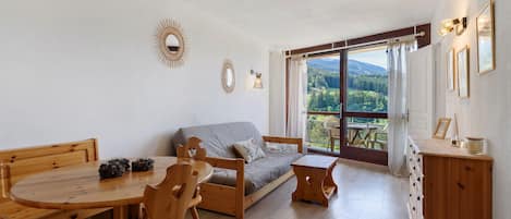 Una habitación acogedora con una mesa de madera, sillas, un sofá y vistas a un bosque a través de un ventanal grande.