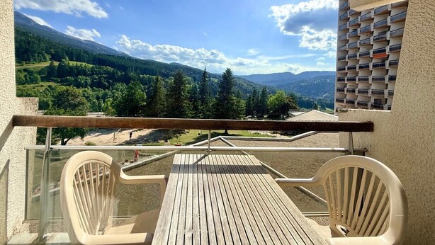 Un balcon avec une table et des chaises en bois, donnant sur un paysage montagneux parsemé d’arbres et sous un ciel dégagé.