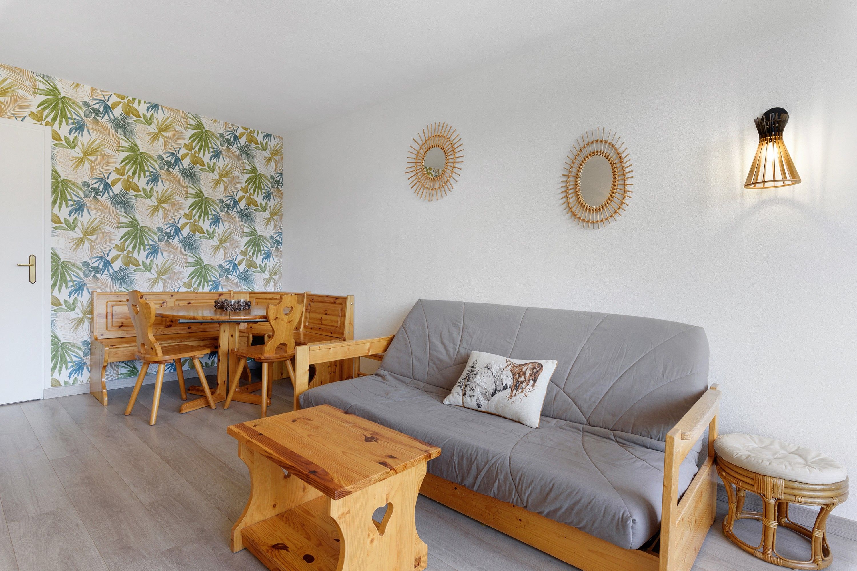 Una sala de estar con un sofá, una mesa de centro de madera y una mesa de comedor con sillas. Hay luminarias fijadas a la pared y una puerta con un papel tapiz de motivos de hojas.