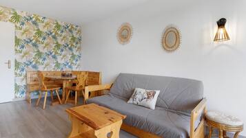 Una sala de estar con un sofá, una mesa de centro de madera y una mesa de comedor con sillas. Hay luminarias fijadas a la pared y una puerta con un papel tapiz de motivos de hojas.