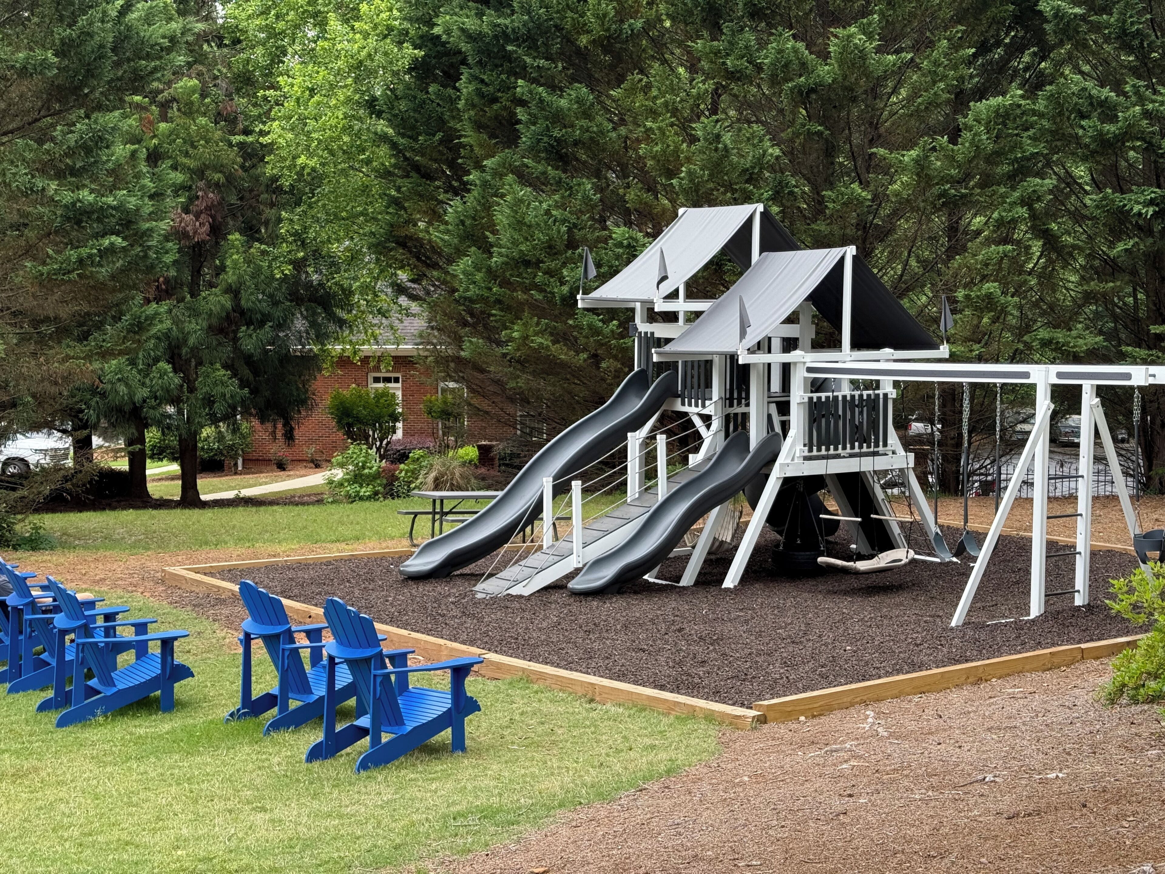 A playground with slides and swings, surrounded by trees and a brick building.