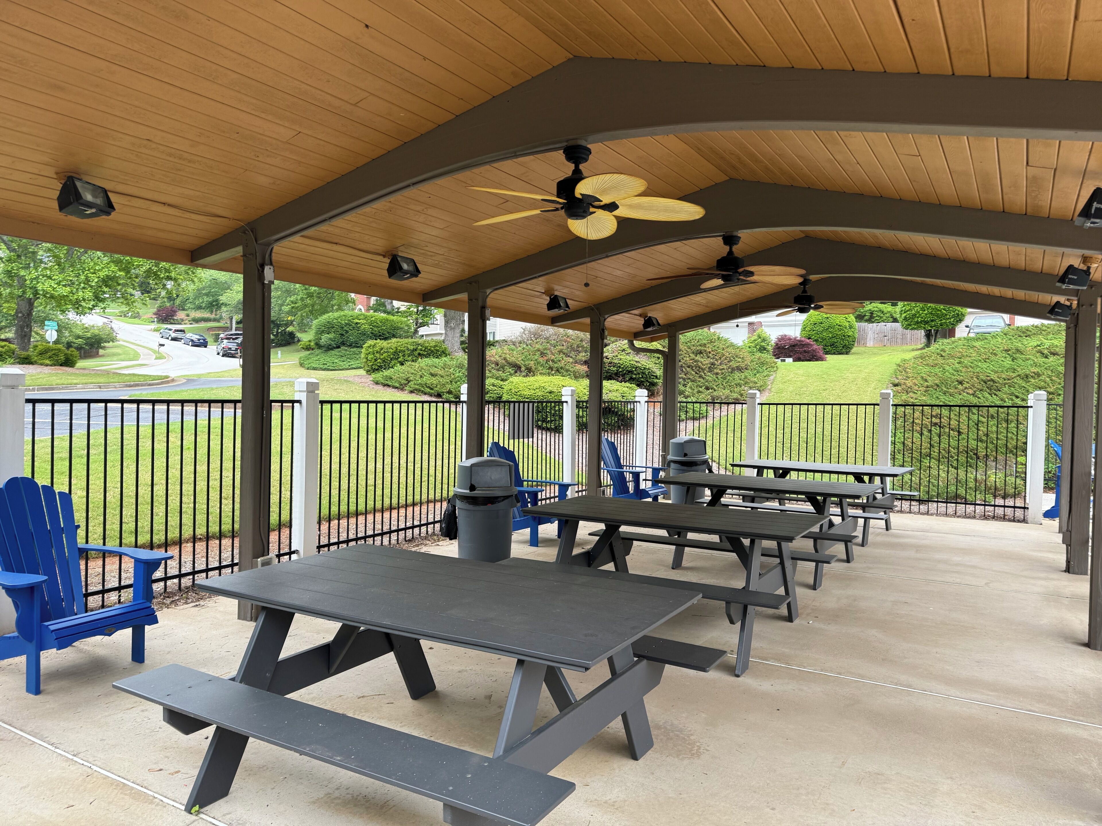 An outdoor covered patio with picnic tables and blue chairs, a trash can, and a view of a green landscape with a road.