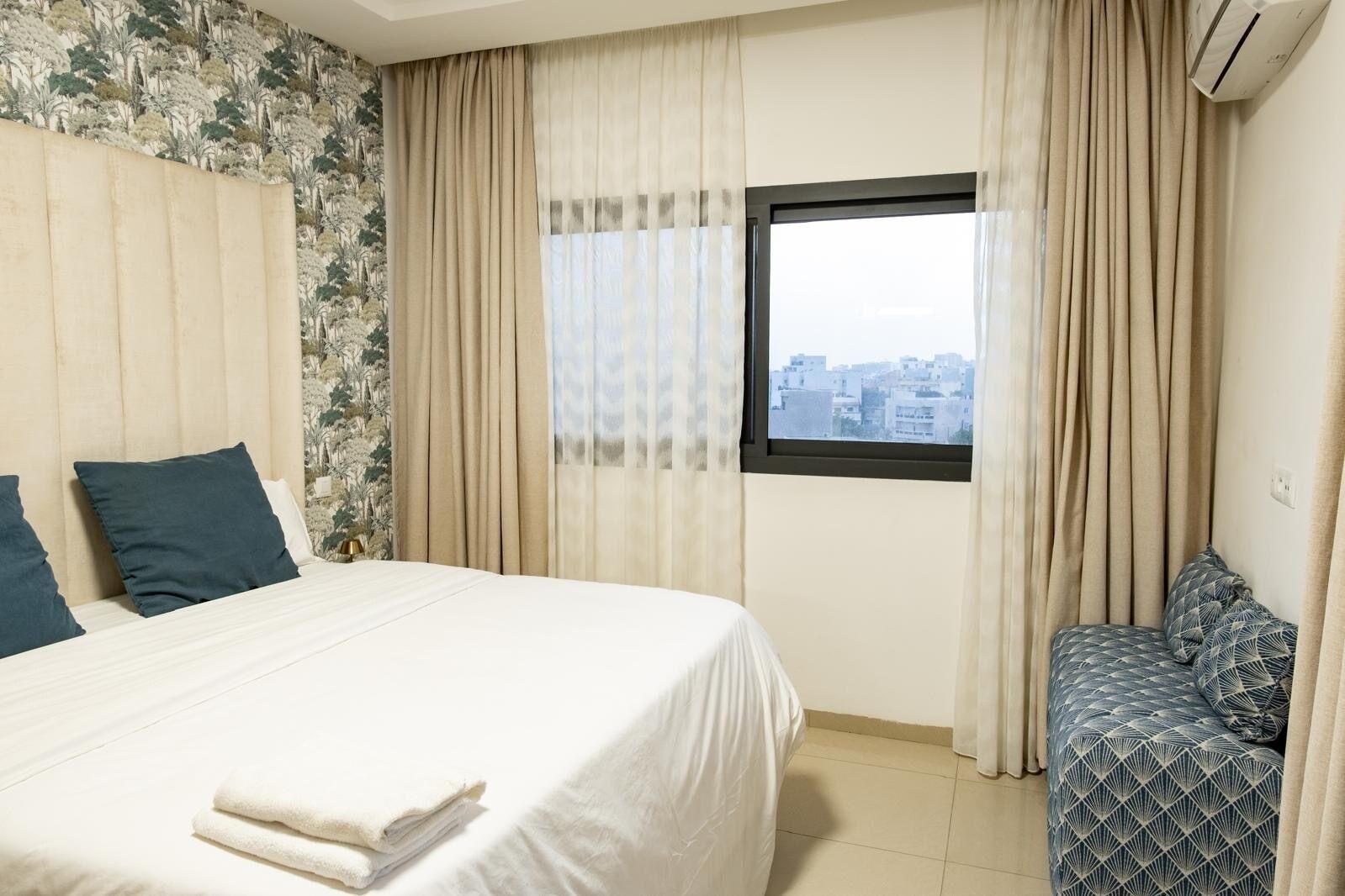 A hotel room with a bed, a bench, and a window with a city view.