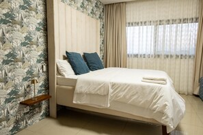 A neatly made bed with white bedding and blue pillows, a bedside table with a lamp, and a window with sheer curtains. - Cozy & Bright 2-Room Apartment in Dakar (Dakar)
