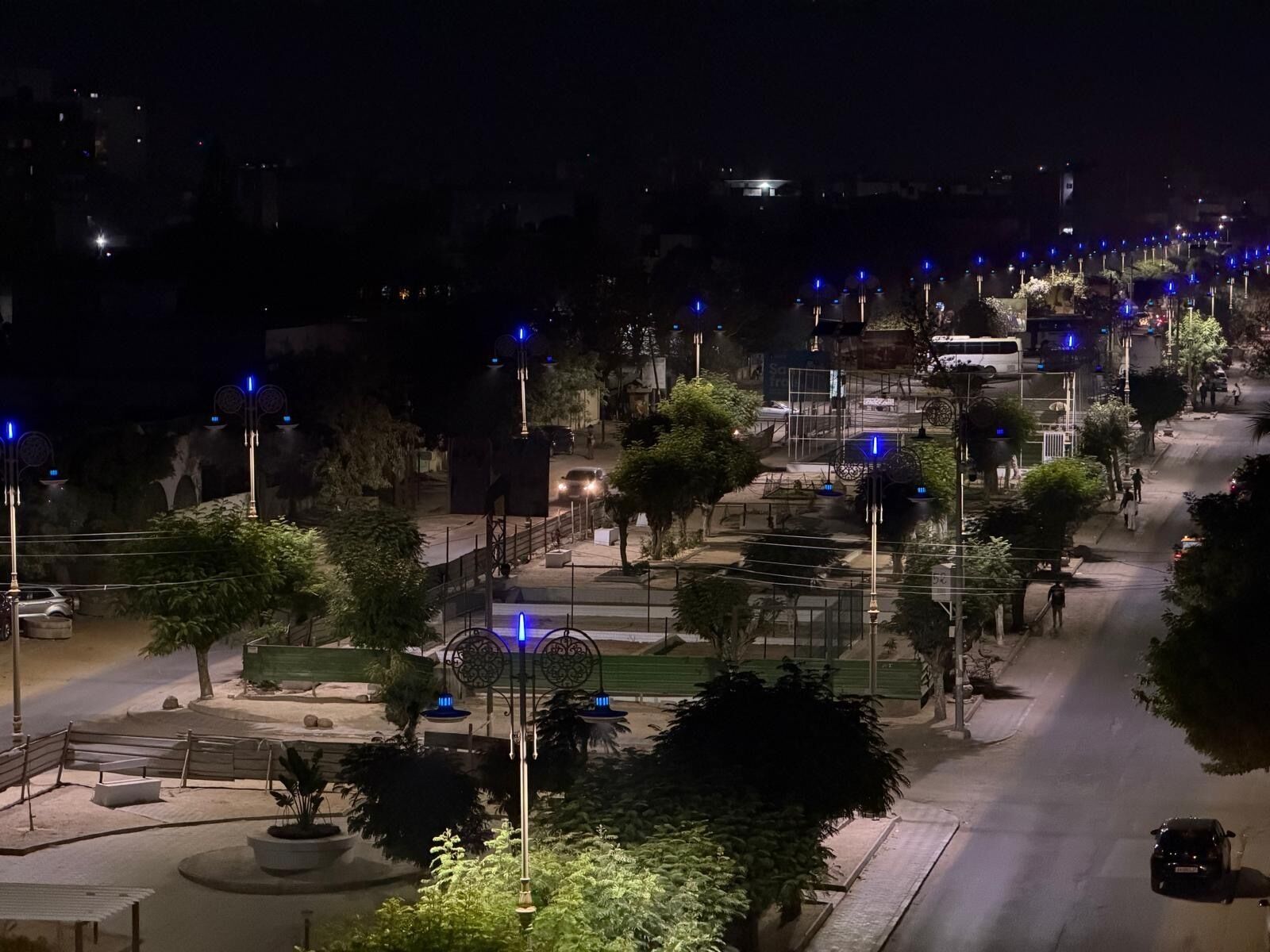 A nighttime street scene with street lamps, trees, and a few parked cars.