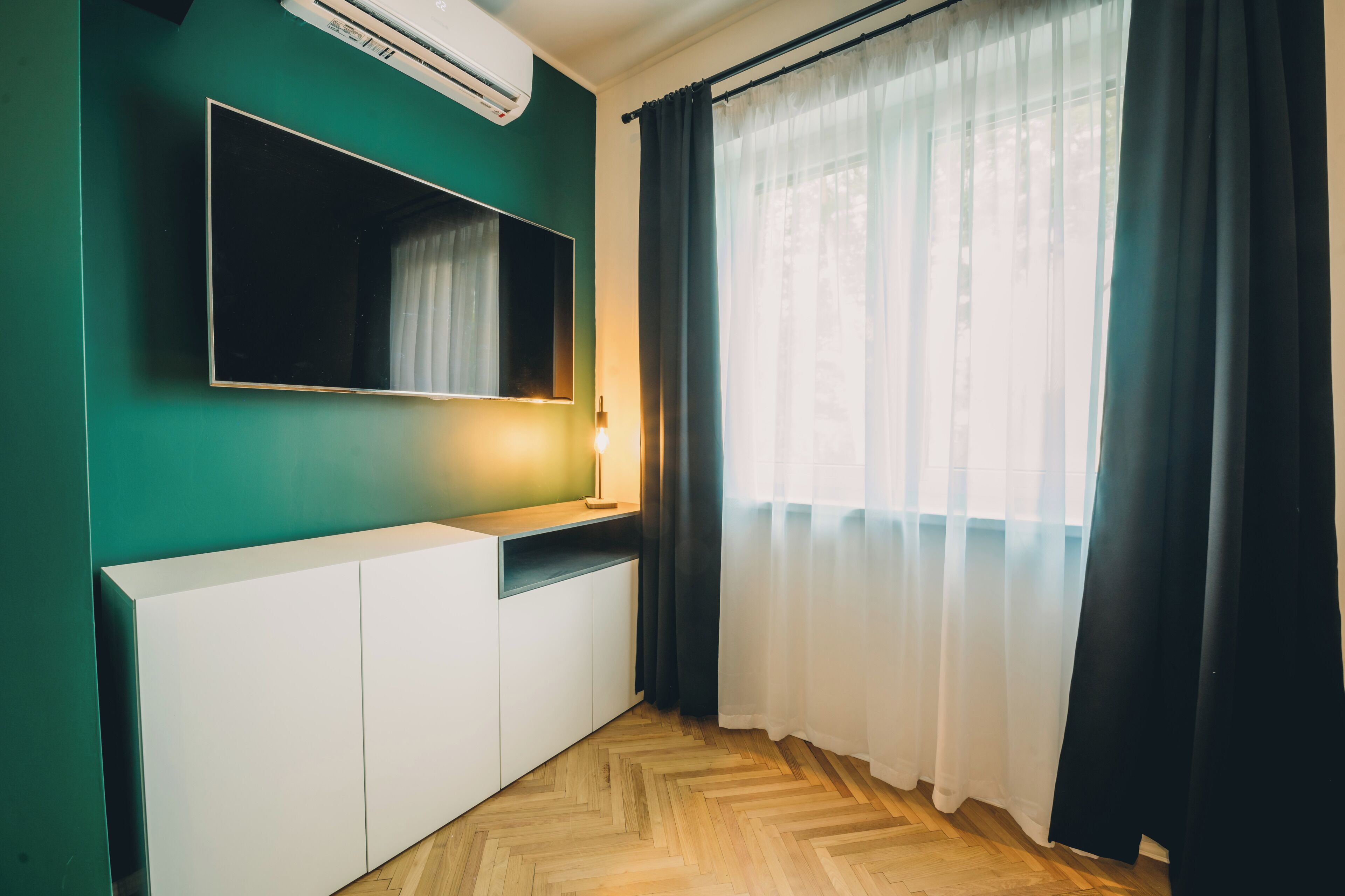 A room with a large flat-screen TV mounted on a teal wall, a white cabinet, a wooden herringbone floor, and a window with sheer white curtains.